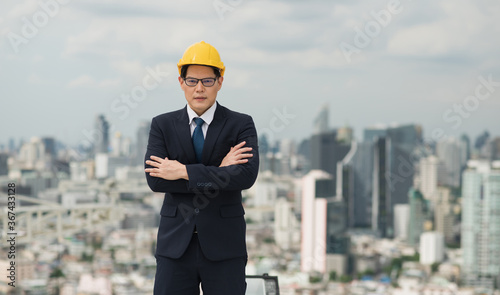 Young handsome asian civil engineer standing with arms crossed wearing helmet yellow hard hat in business suite on building urban cities background.