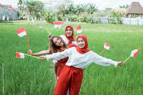 three Asian girls smile posing hold a small flag standing in the middle of rice fields while wearing the Indonesian flag attribute photo