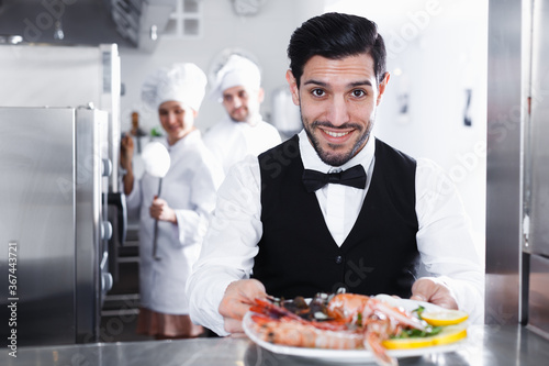 Smiling bearded waiter holding seafood dish in kitchen of fish restaurant