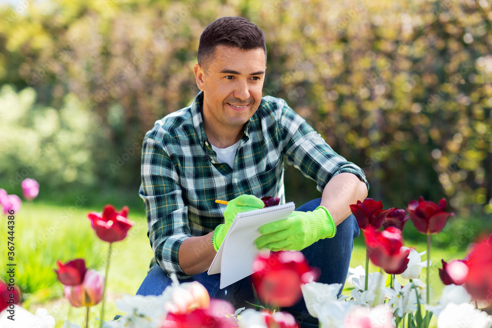 gardening and people concept - happy smiling middle-aged man writing to notebook and taking care of flowers at summer garden gardening and people concept - happy smiling middle-aged man writing to notebook and taking care of flowers at summer garden