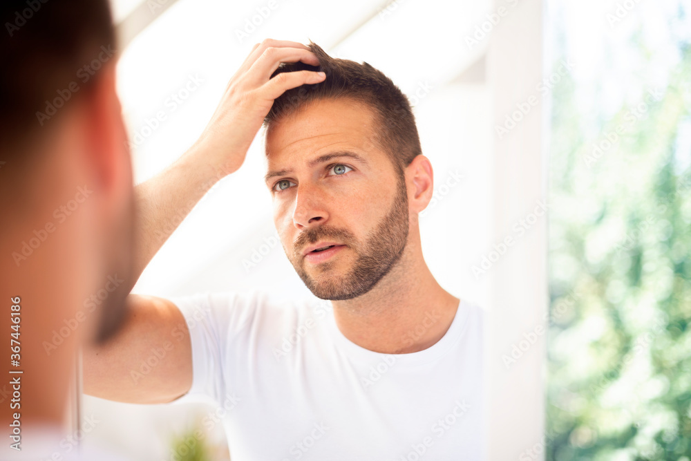 Handsome young man standing in front of bathroom mirror and looking his face