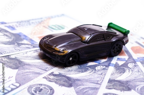 Black toy car on a heap of dollar bills on a white and black background. A sports car with a green spoiler  a wing on dollars in a close-up top and side view. Rear and front car. Selling  buying