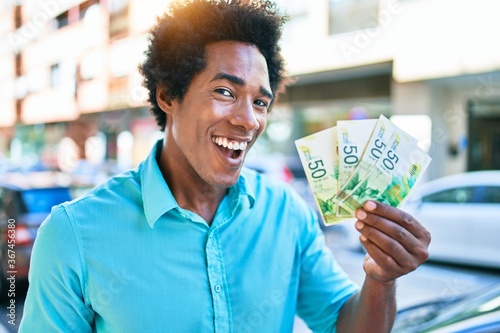 Young african american man smiling happy. Standing with smile on face holding israeli shekels banknotes at town street. photo
