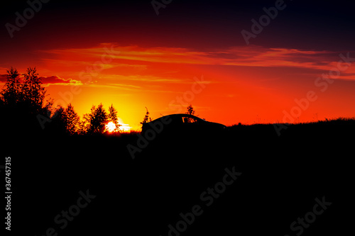 Summer sunset in a field in the sky of black bright colorful red orange color with clouds. The sun sets over the horizon against the background of the landscape of silhouettes of trees grass car