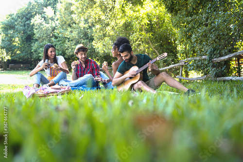 Group of young people having fun having a picnic in the countryside. Mixed race people drinking wine, eating sandwiches and playing guitar outdoors. Healthy lifestyle concept.