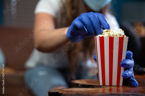 Woman wearing safety mask and gloves for Covid-19, coronavirus and quarantine watching television at home with red and white striped cupboard bucket with popcorn. Cinema and health concept