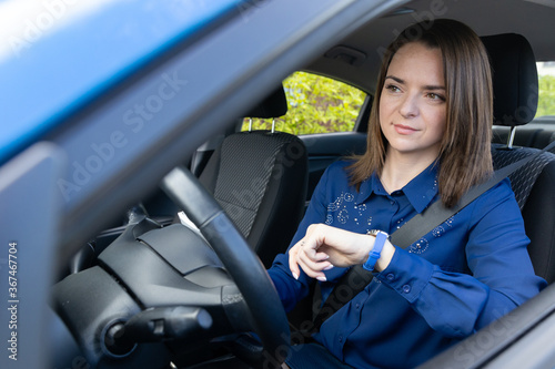 A business-style girl sits behind the wheel of a car and looks at her watch