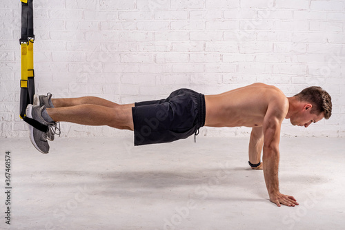 Muscular young man doing exercises with trx straps, near a white brick wall.