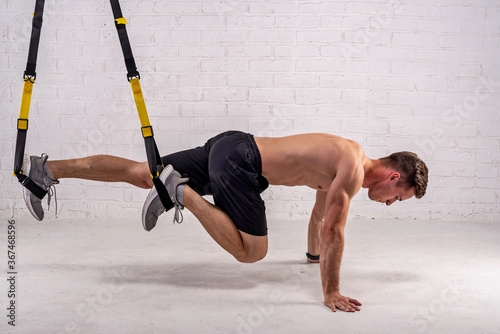 Muscular young man doing exercises with trx straps, near a white brick wall.