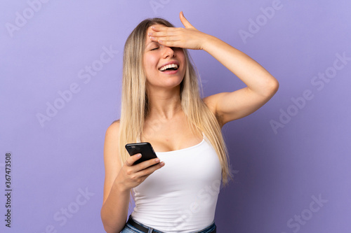 Young blonde woman using mobile phone isolated on purple background laughing