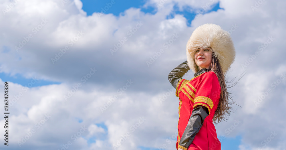 A young girl in a national Caucasian costume (Lezginka) and in a white fur hat stands against a blue cloudy sky. Summer Adventures. A young girl in a national Caucasian costume (Lezginka) and in a white fur hat stands against a blue cloudy sky. Summer Adventures.