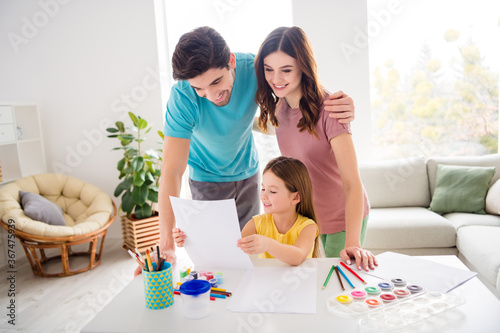 Photo of positive cheerful three people small kid girl sit table show mommy daddy beautiful painted picture paper in house indoors