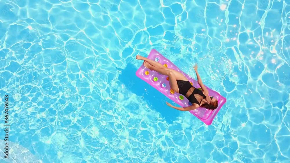 Fitted pretty girl in bikini chilling on inflatable pink mattress in swimming pool. Shaped woman in swimsuit tanning. Female relaxing on float in blue water at luxury resort. Aerial, view from above.