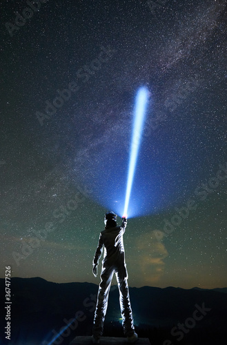 Back view of space traveler in space suit and helmet illuminating beautiful starry sky with flashlight while standing under magical night sky with Milky Way. Concept of light, astronomy, cosmonautics