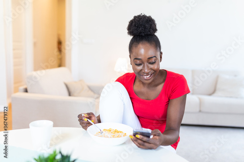 Beautiful young arfican american woman texting on smart phone at home. Cheerful lady eating corn flakes, drinking morning coffee and looking at her mobile phone
