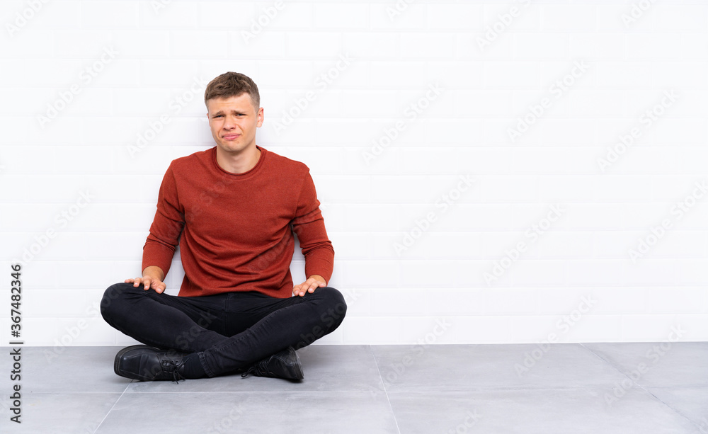 Young handsome man sitting on the floor having doubts and with confuse face expression Young handsome man sitting on the floor having doubts and with confuse face expression