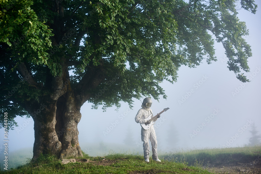 Space traveler playing melody on guitar near large tree with green leaves. Astronaut guitarist with musical instrument wearing white space suit and helmet while standing in misty valley with big tree. Space traveler playing melody on guitar near large tree with green leaves. Astronaut guitarist with musical instrument wearing white space suit and helmet while standing in misty valley with big tree.