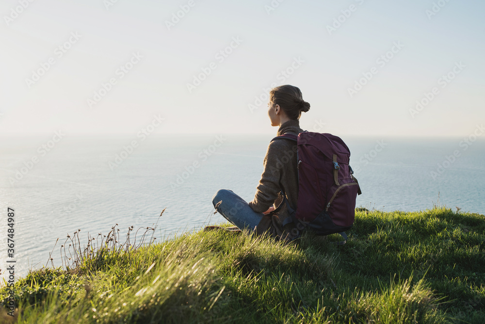 Young woman traveler with backpack sitting and looking at sea over beautiful cliffs background, Travel, active lifestyle and summer holiday concept
