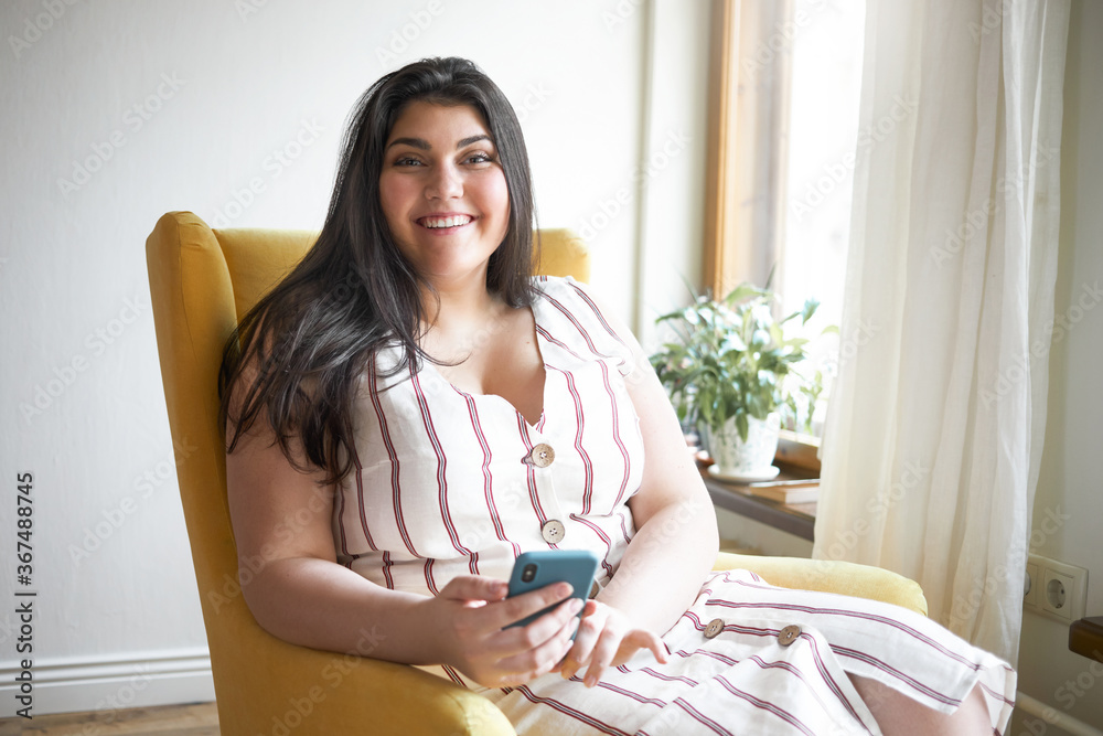 Cheerful young overweight Caucasian woman with curvy body looking at camera with broad happy smile holding smart phone, using high speed internet connection, chatting with friends online via messenger Cheerful young overweight Caucasian woman with curvy body looking at camera with broad happy smile holding smart phone, using high speed internet connection, chatting with friends online via messenger