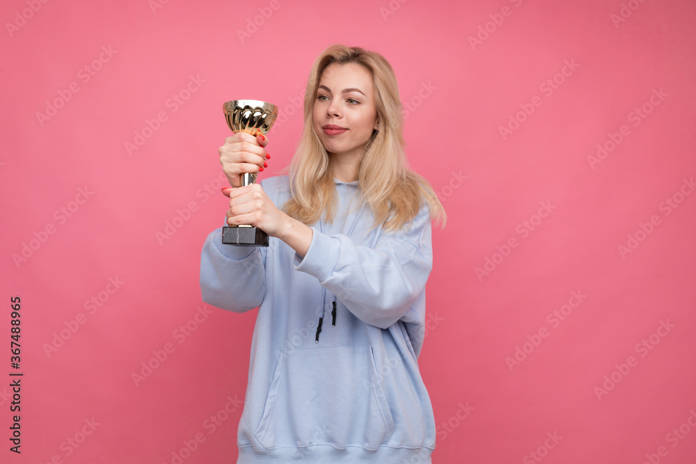 A young girl in a fashionable hoodie holds a gold cup on a pink background. A young girl in a fashionable hoodie holds a gold cup on a pink background.