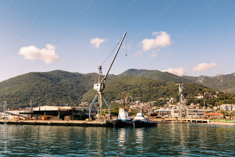 Cranes at cargo terminal in Kotor bay. Montenegro docking station with cranes and tug boats. Mountains, town, blue sky as background.