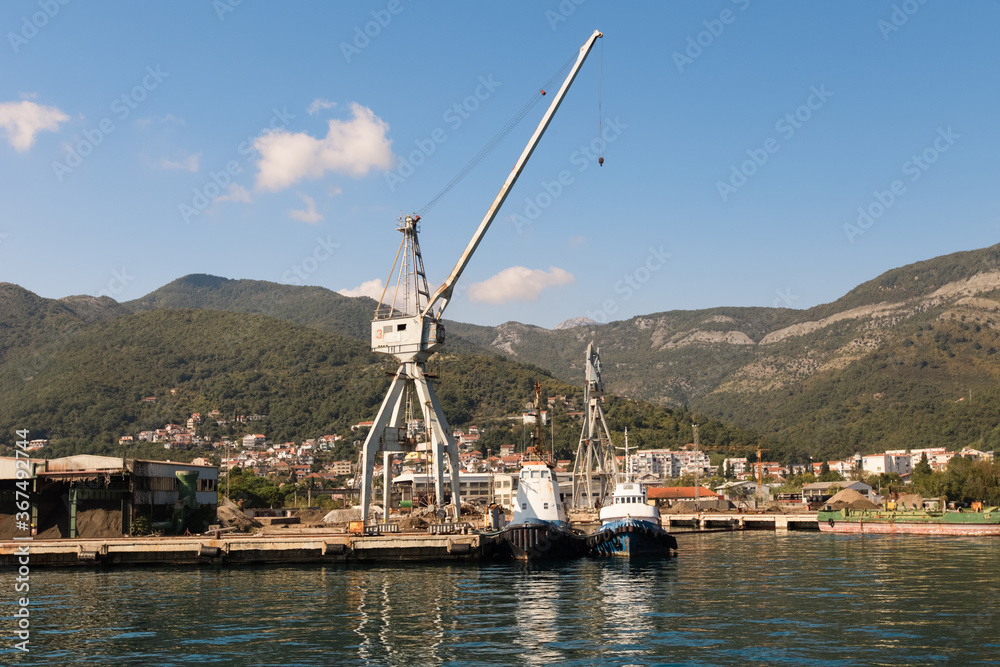 Cranes at cargo terminal in Kotor bay. Montenegro docking station with cranes and tug boats. Mountains, town, blue sky as background.