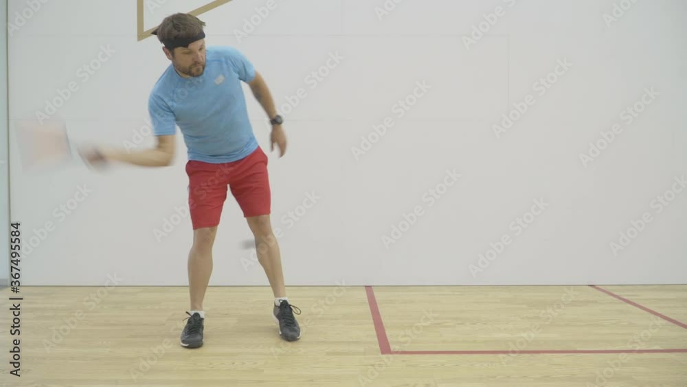 Wide shot of strong Caucasian mid-adult sportsman training in gym. Portrait of brunette bearded athletic man exercising indoors. Confident concentrated guy playing squash racket and ball sport. Wide shot of strong Caucasian mid-adult sportsman training in gym. Portrait of brunette bearded athletic man exercising indoors. Confident concentrated guy playing squash racket and ball sport.