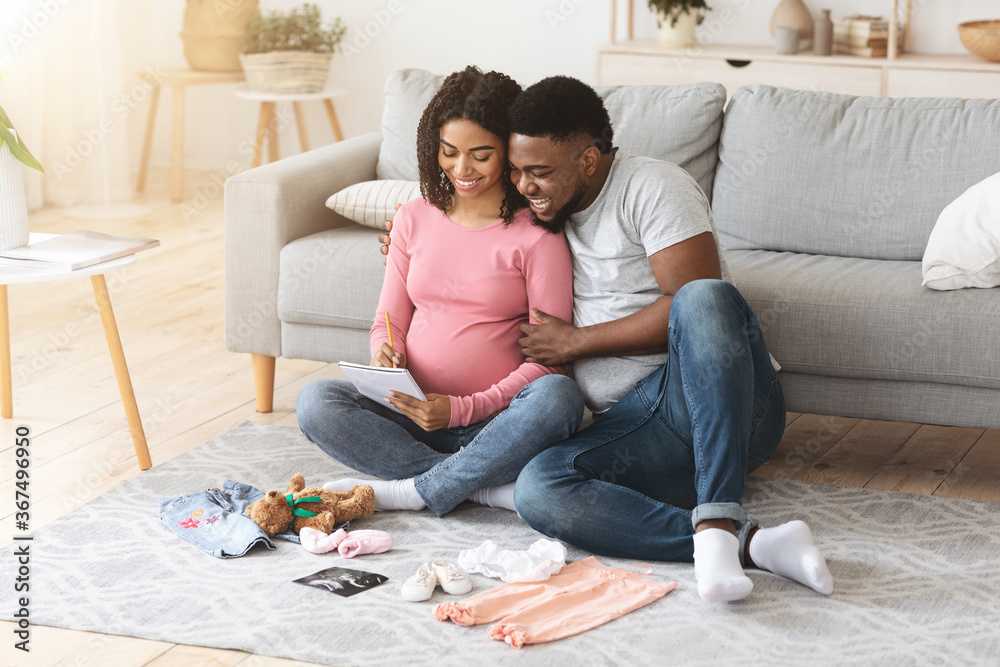 Fototapeta premium Cheerful pregnant black family preparing baby clothes and making list
