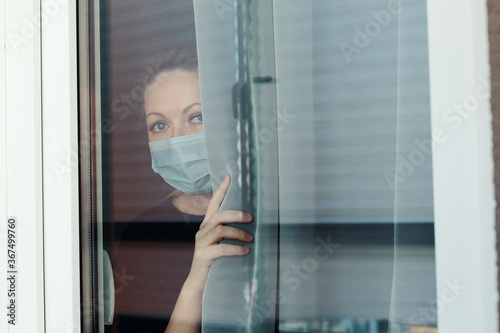 Quarantine woman in medical mask on face looking through the window.