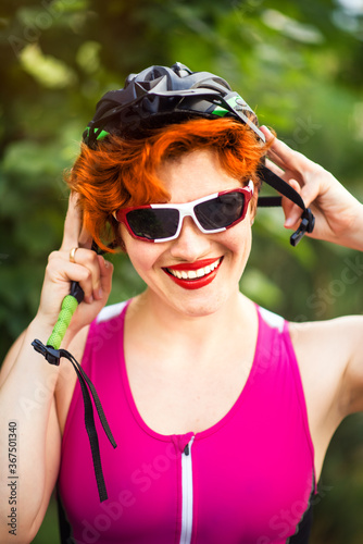 Vertical portrait of sexy red haired smiling woman cyclist in pink bike suit and sun glasses wearing bike helmet. Active summer sport for woman. Outdoor sport activity portrait.