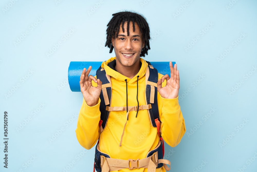 Young mountaineer african american man with a big backpack isolated on a blue background showing an ok sign with fingers Young mountaineer african american man with a big backpack isolated on a blue background showing an ok sign with fingers