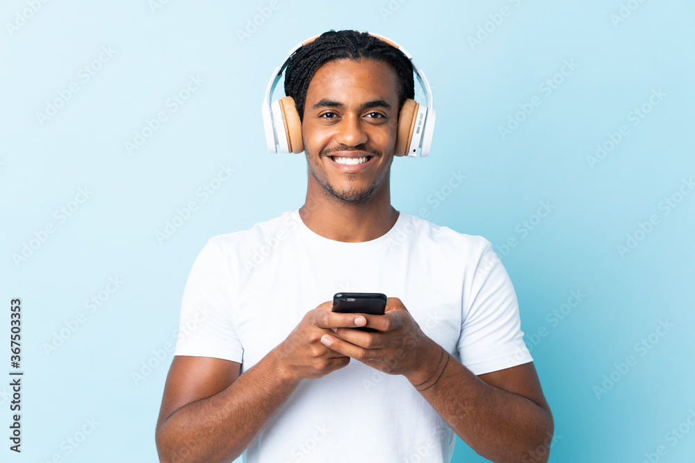 Young African American man with braids isolated on blue background listening music with a mobile and looking front Young African American man with braids isolated on blue background listening music with a mobile and looking front