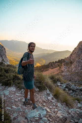 Young spanish man standing while looking at camera making a "cool" sign during a hike path in Serra de Tramuntana at golden hour with the village of Soller at the backgorund tinted by golden light