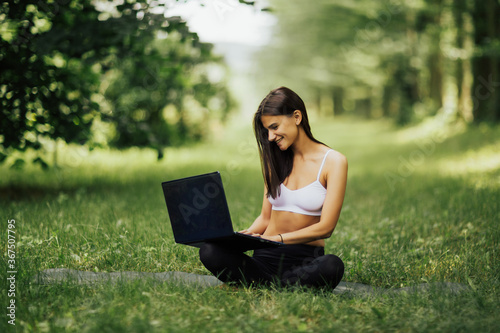 Smiling young fitness girl using laptop computer while sitting on the yoga mat in park. Beautiful young woman practicing yoga in garden outdoors following guide of online trainer on laptop. © eduard