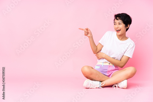 Young Vietnamese woman with short hair sitting on the floor over isolated pink background pointing finger to the side and presenting a product