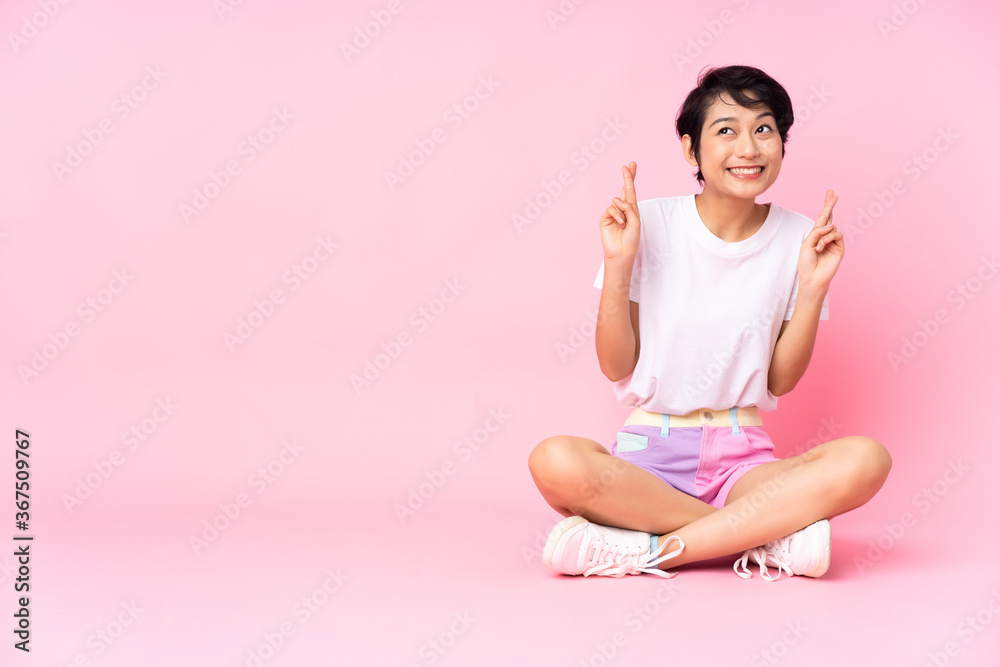 Young Vietnamese woman with short hair sitting on the floor over isolated pink background with fingers crossing Young Vietnamese woman with short hair sitting on the floor over isolated pink background with fingers crossing