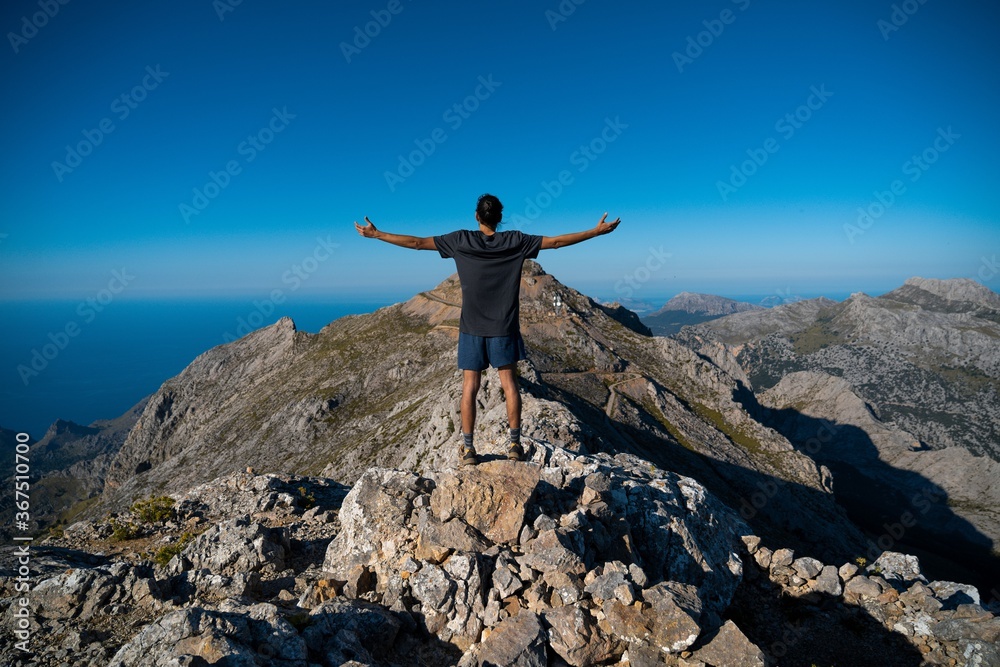 A man stands on a rock with his arms raised during a trekking on a mountain looking at the stunning views of Serra de Tramuntana (Mallorca, Spain)