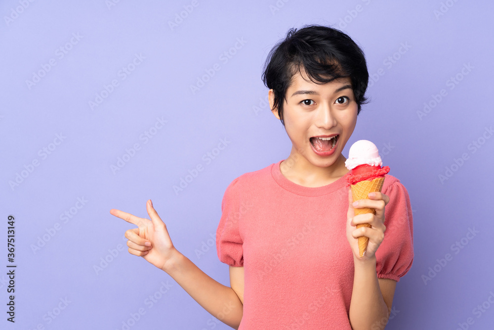 Young Vietnamese woman with short hair holding a cornet ice cream over isolated purple background surprised and pointing finger to the side Young Vietnamese woman with short hair holding a cornet ice cream over isolated purple background surprised and pointing finger to the side