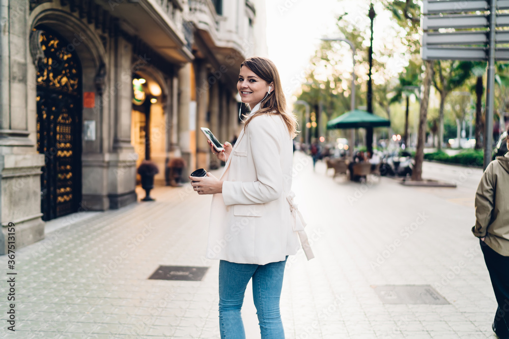 Half length of millennial user in headphones connecting to 4g internet for listening music songs during travel walking in city, happy female blogger with mobile phone and coffee to go enjoying pastime