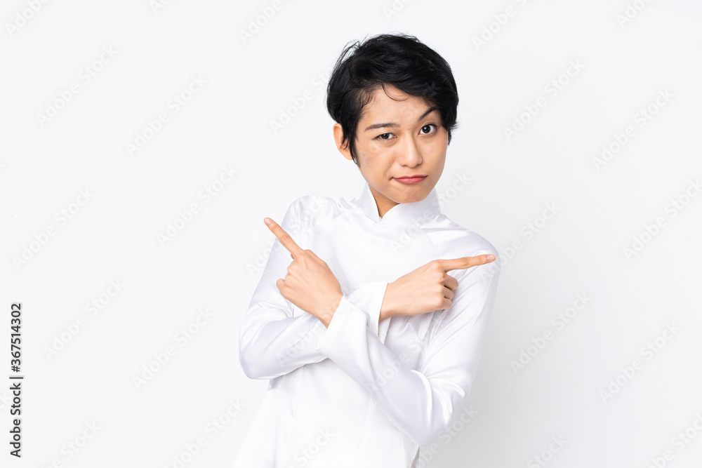 Young Vietnamese woman with short hair wearing a traditional dress over isolated white background pointing to the laterals having doubts