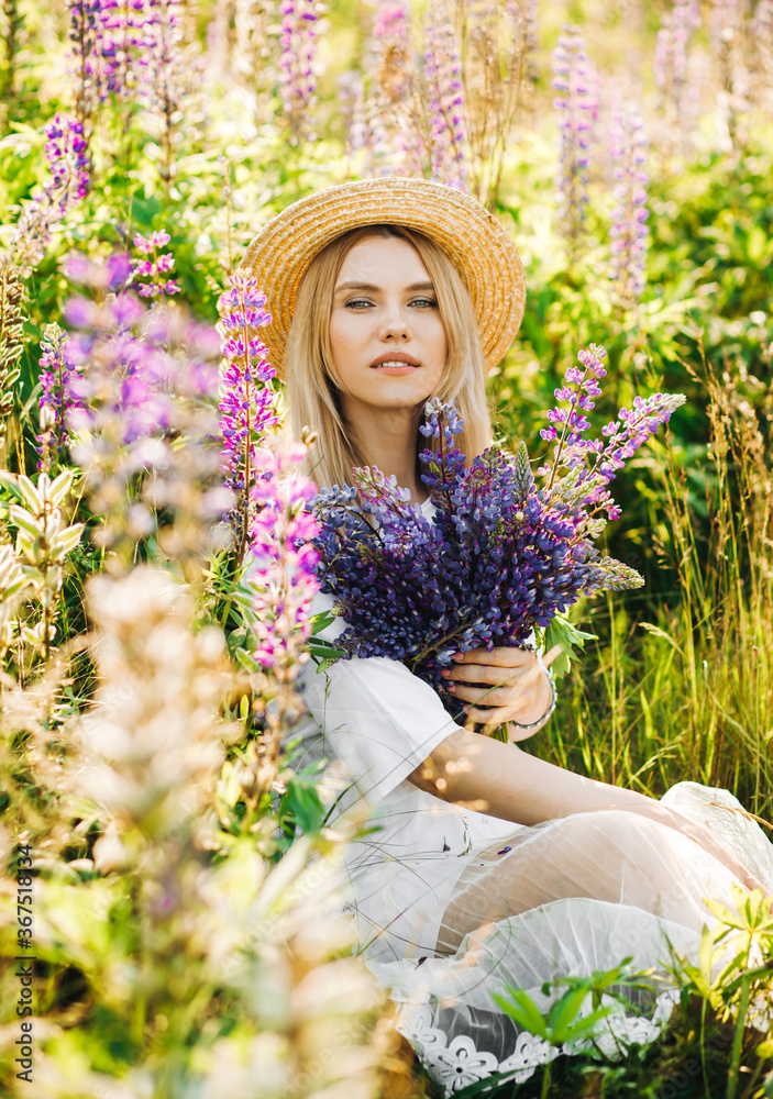Charming woman in a straw hat sits in a field holding a bouquet of lupines in her hands.