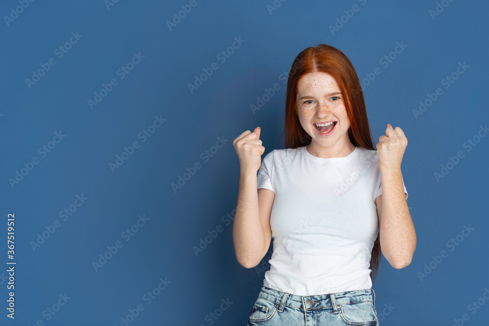 Winner, celebrating. Caucasian young girl's portrait on blue background. Beautiful female redhair model with cute freckles. Concept of human emotions, facial expression, sales, ad, youth culture.