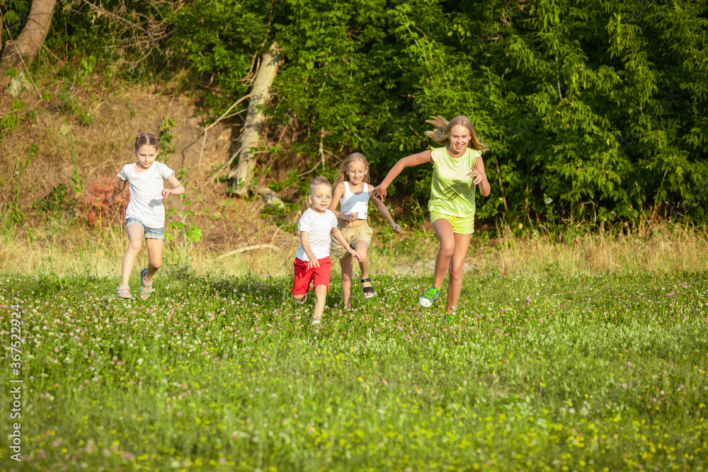 Fototapeta premium Kids, children running on meadow in summer's sunlight. Look happy, cheerful with sincere bright emotions. Cute caucasian boys and girls. Concept of childhood, happiness, movement, family and summer.