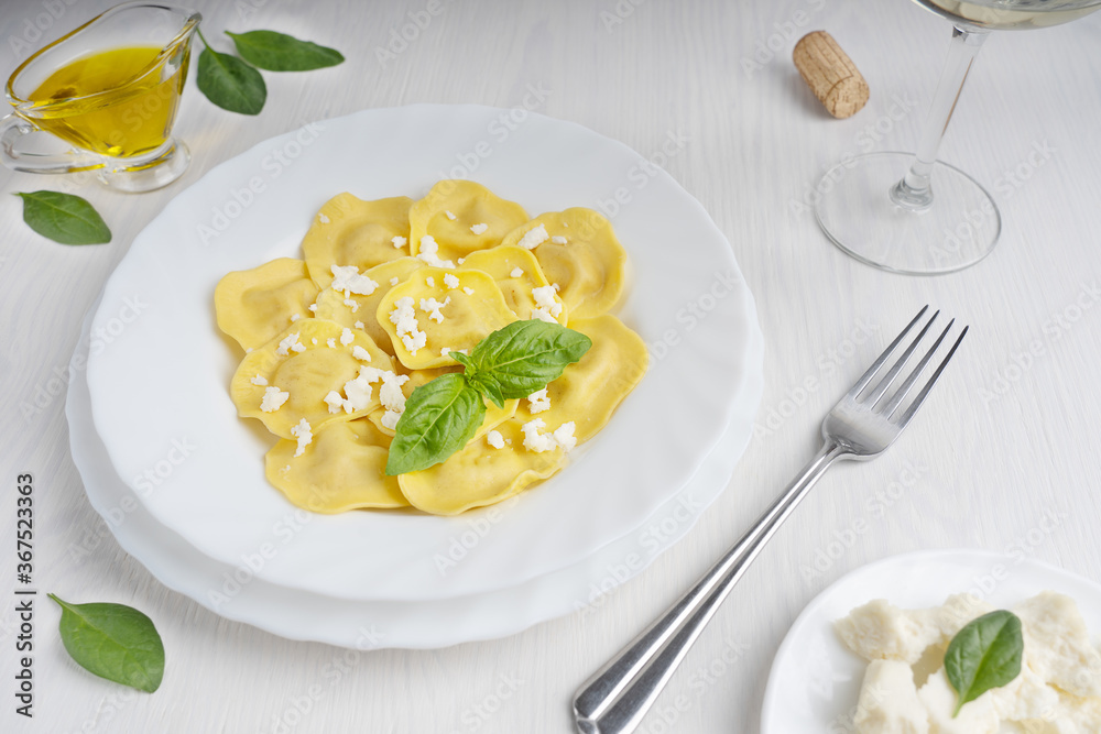 Italian ravioli pasta in a round plate decorated by ricotta cheese and basil leaves with a glass of white wine and olive oil gravy boat standing on white wooden background ready for eating