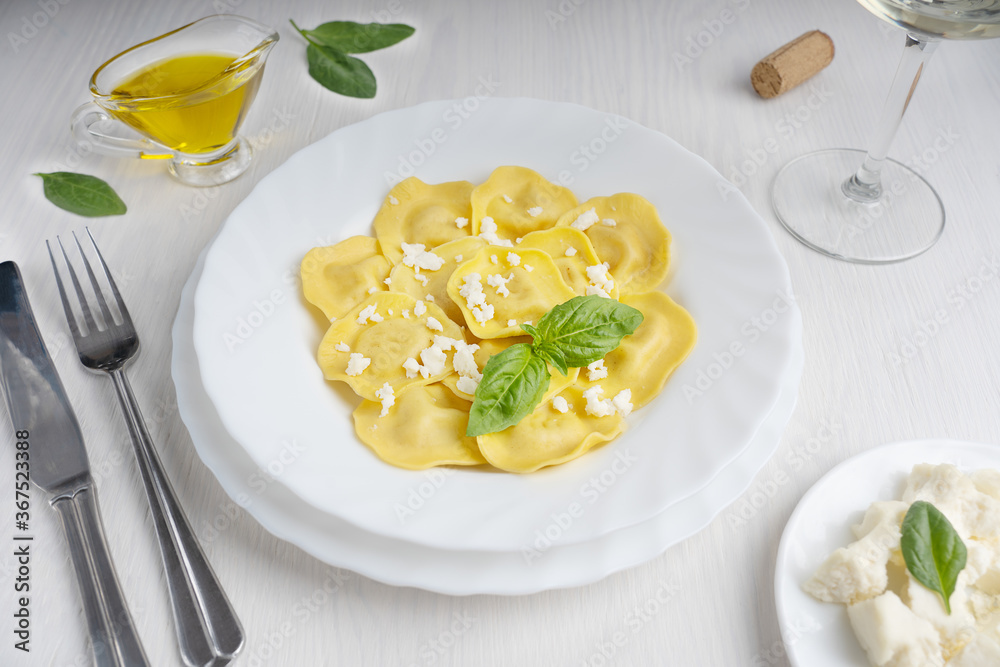 Italian ravioli pasta in a round plate decorated by fresh basil leaves surrounded by olive oil gravy boat, knife and fork and glass of wine ready for eating on white wooden background at restaurant Italian ravioli pasta in a round plate decorated by fresh basil leaves surrounded by olive oil gravy boat, knife and fork and glass of wine ready for eating on white wooden background at restaurant
