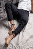 Cropped shot of a man in white t-shirt and black trousers is lying on the gray bed sheets. Man's legs are shackled by chrome steel ankle cuffs with chain and bronze padlocks.