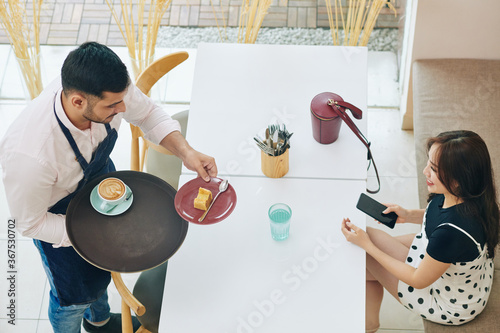 Waiter bringing piece of delicious cake and cup of cappuccino to pretty young woman at cafe table
