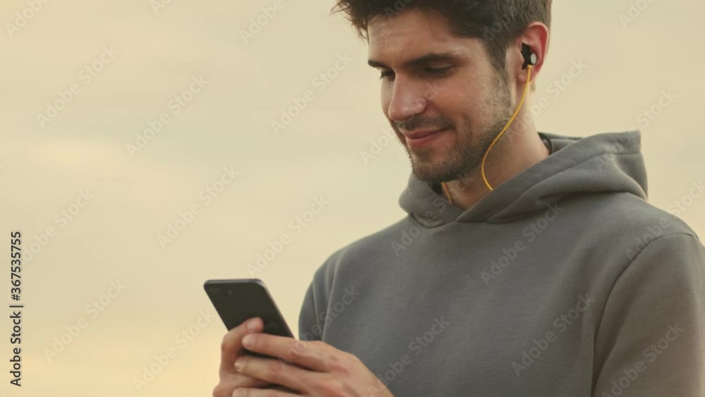 A pleased young sportsman with earphones is using his smartphone while walking outside in the morning A pleased young sportsman with earphones is using his smartphone while walking outside in the morning