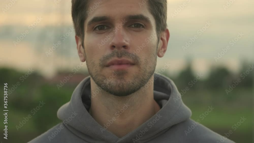 A close-up view of a young sportsman in hoodie is opening his eyes and looking to the camera while walking outside