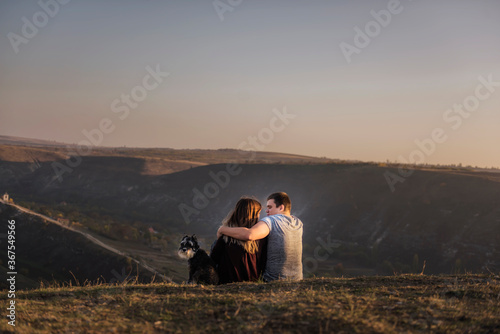 A loving couple of travelers with a Schnauzer dog in their arms are sitting on a rock, hugging each other, looking at the opening sunset panorama view of the river. A trip of a happy family with pets 